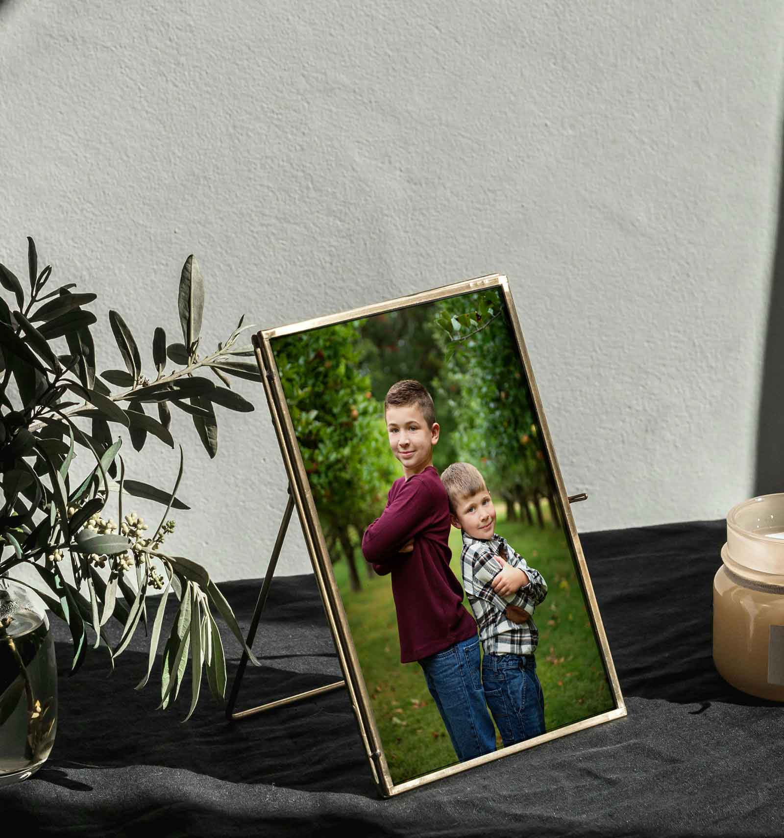A framed photo of two boys standing back to back outdoors, placed on a black table next to a plant and a candle.