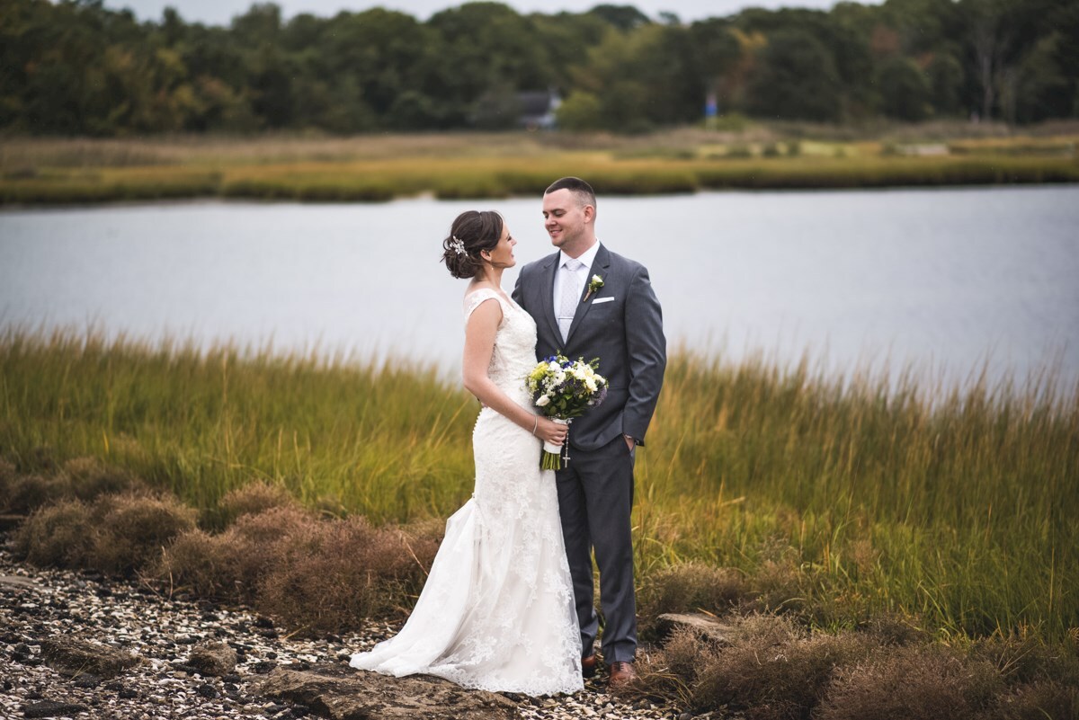 A bride and groom stand together outdoors by a lake, facing each other. The bride holds a bouquet, and both are dressed formally, with greenery and water in the background.