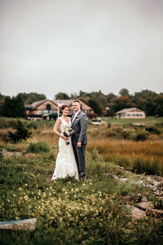 harbor-lights-wedding-015 A bride and groom stand together outdoors on a grassy field, with houses and trees in the background under a cloudy sky.
