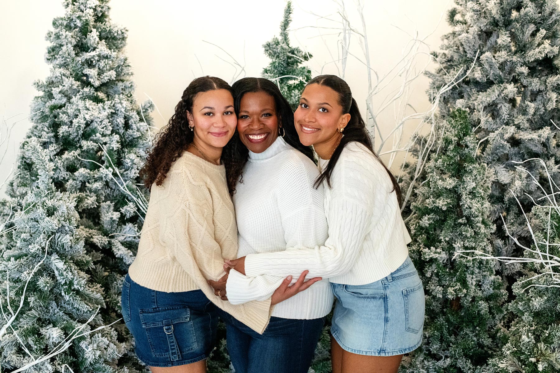 Three women stand closely together, smiling, in front of snow-covered artificial pine trees. They are wearing light sweaters and denim skirts or jeans.