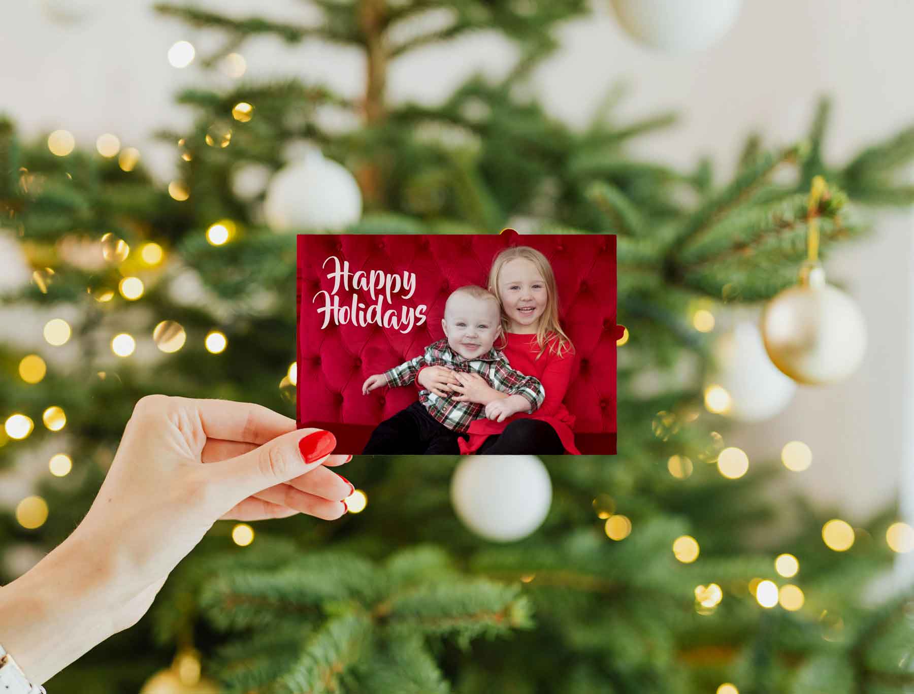 A hand with red nail polish holds a holiday card showing two smiling children in festive clothing, in front of a decorated Christmas tree with white ornaments and lights.
