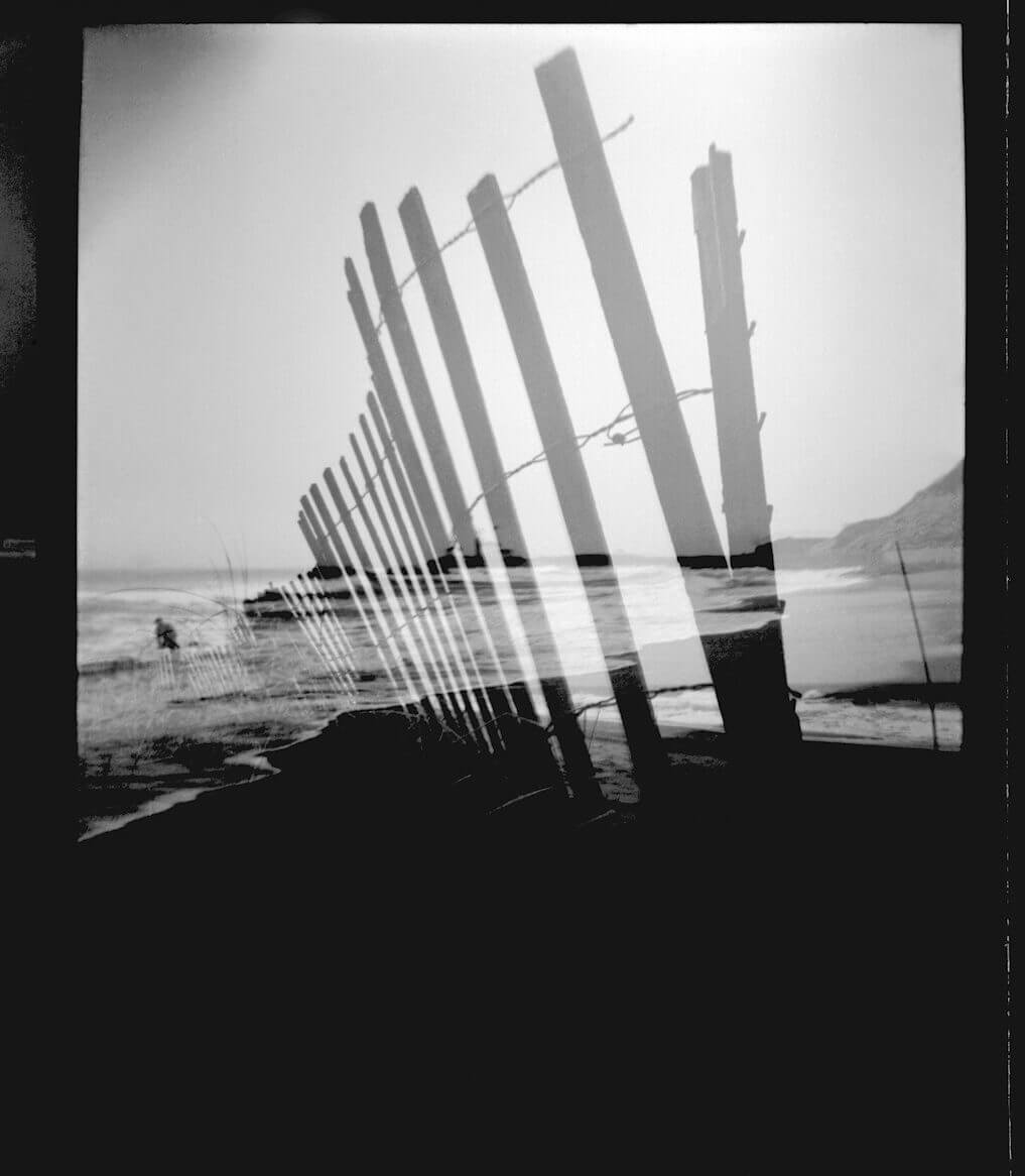 jbd-beachfence-bw Black and white photo showing a weathered wooden fence along a beach, with a double exposure effect creating a layered, distorted appearance.