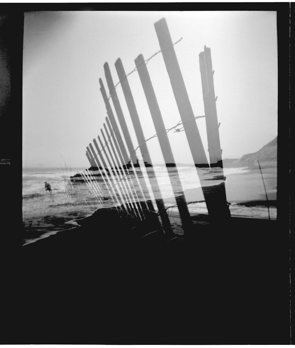 jbd-beachfence-bw Black and white photo showing a weathered wooden fence along a beach, with a double exposure effect creating a layered, distorted appearance.