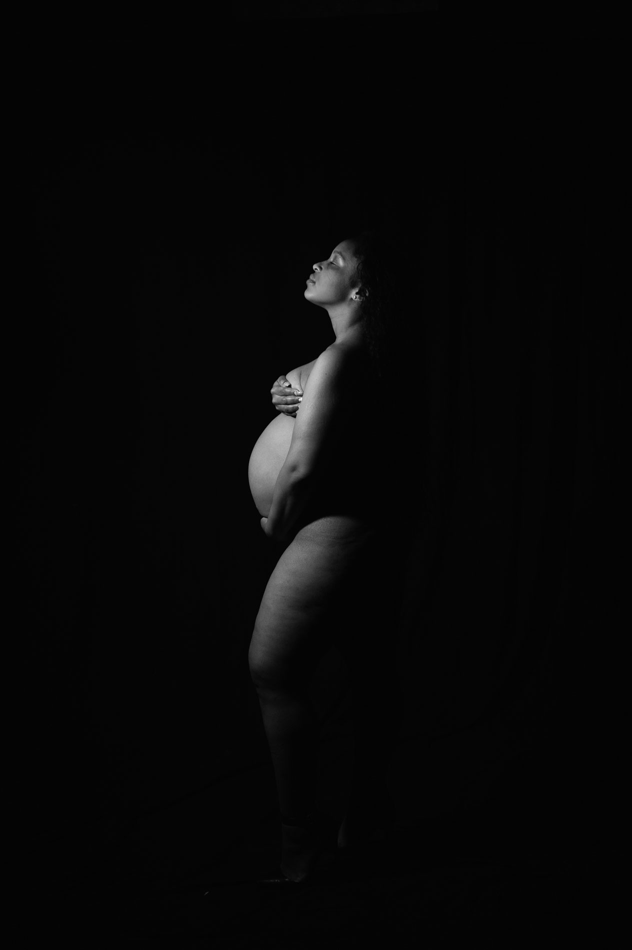 maternity-photography-black-and-white A nude pregnant person stands in profile against a dark background, looking upward with hands crossed over their chest, softly lit by a focused light source.