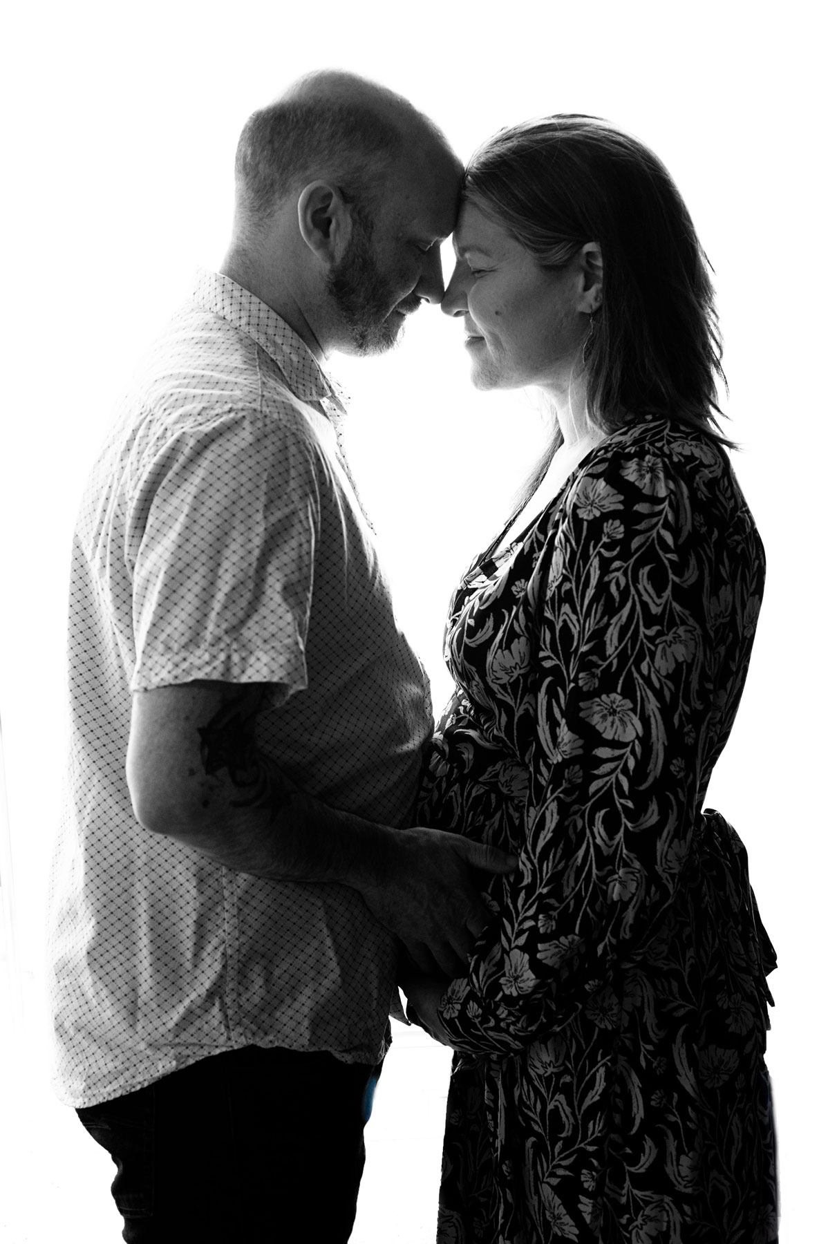 A man and a woman stand face-to-face, touching foreheads and smiling, with the woman holding her pregnant belly.