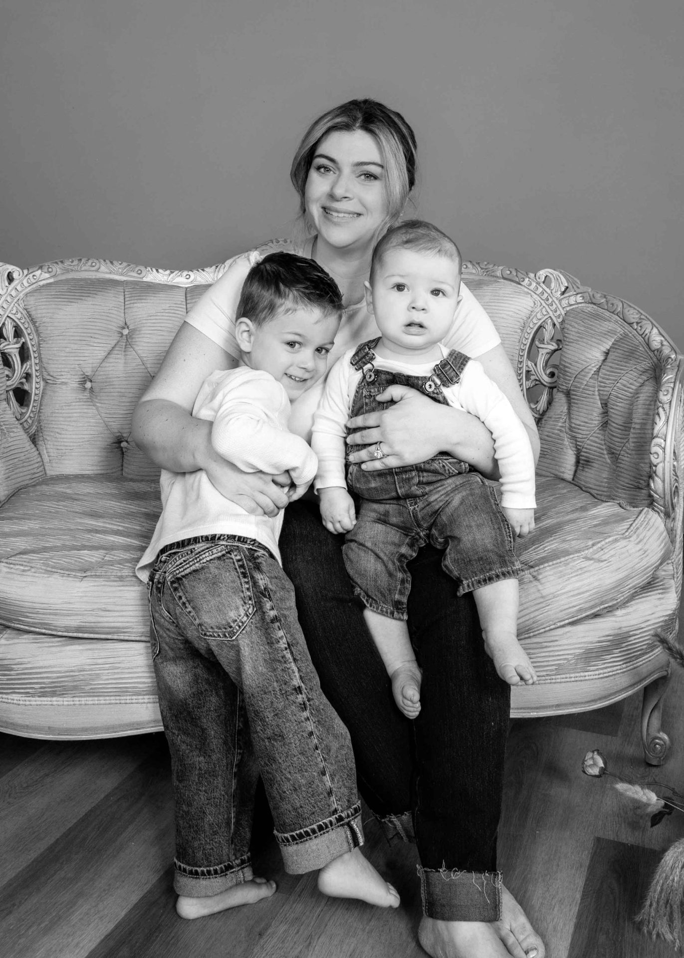 mothers-day-woman-and-two-young-children-on-couch-bw A woman sits barefoot on a vintage sofa, holding a baby on her lap and hugging a young child, all dressed casually, against a plain background.