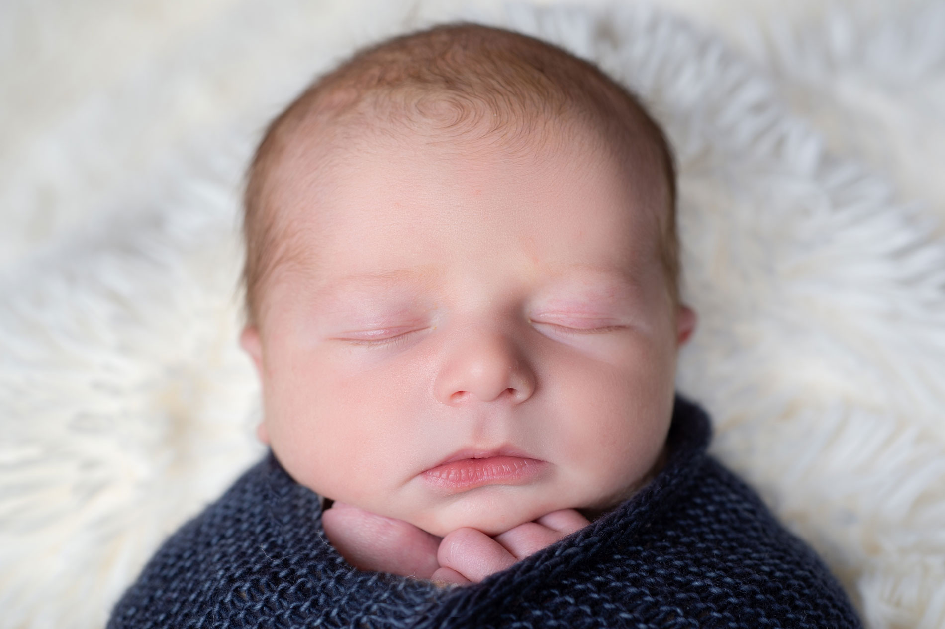 newborn-photography-infant-in-blue-blanket A newborn baby with closed eyes is swaddled in a dark blue blanket, resting on a soft, white textured surface.