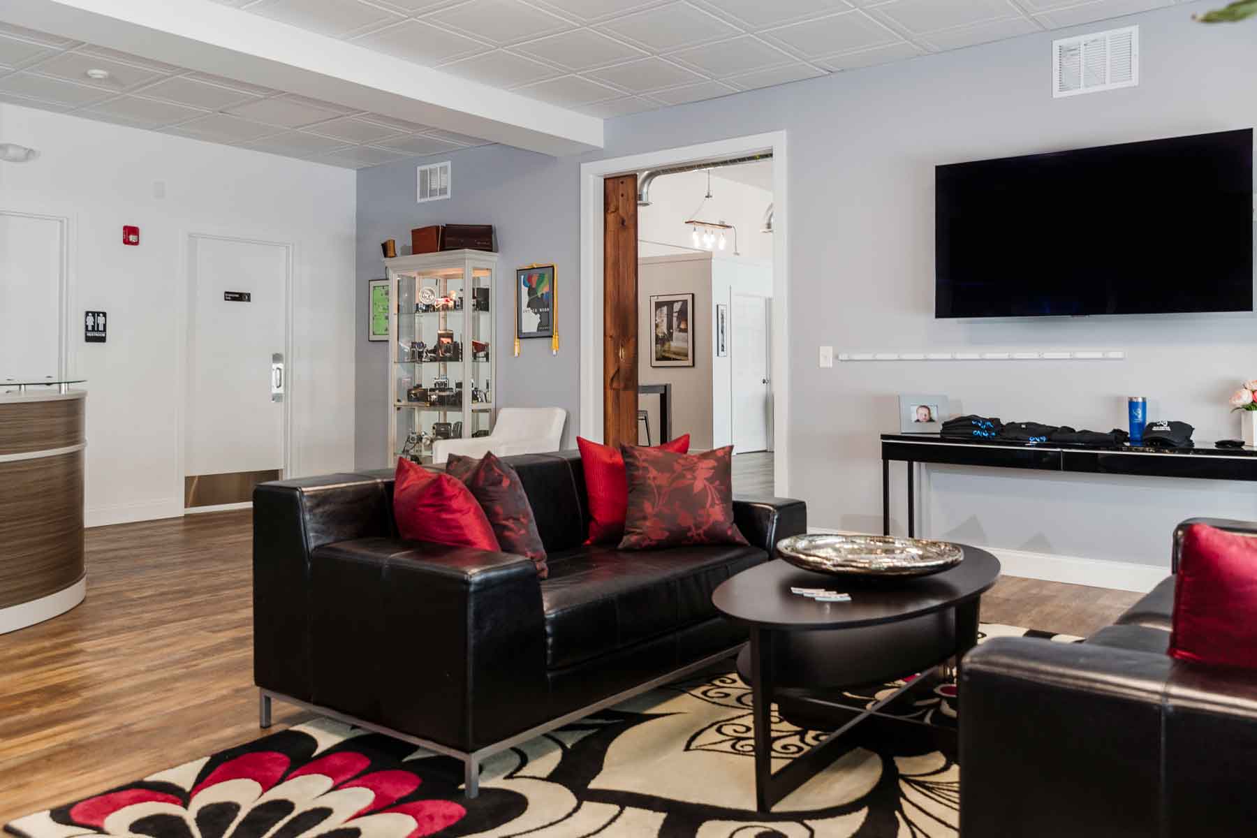 Modern waiting room with black leather sofas, red accent pillows, a round coffee table, a large wall-mounted TV, and a display case in the background.