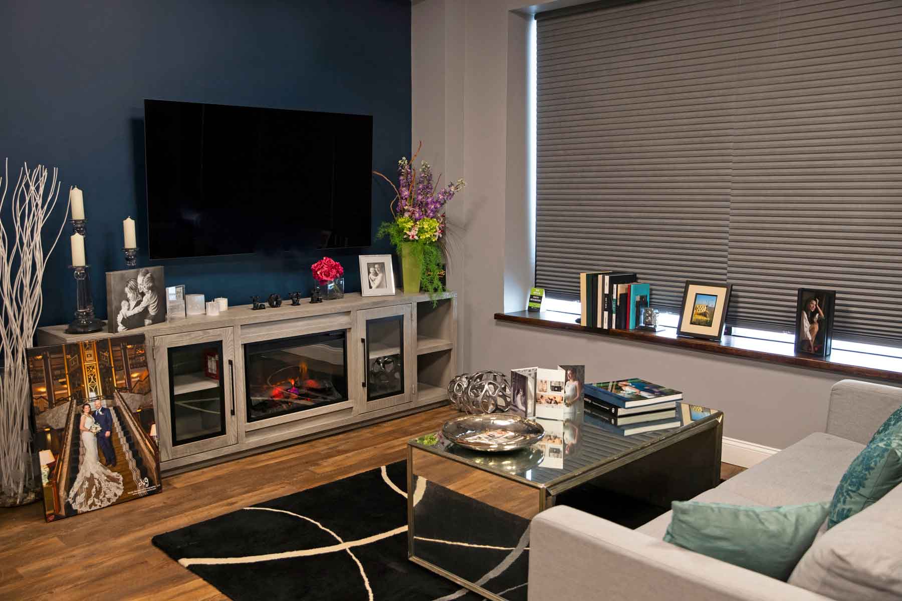 A modern living room with a wall-mounted TV, a decorated mantel, framed photos, a gray sofa, a glass coffee table, and a window with closed blinds.