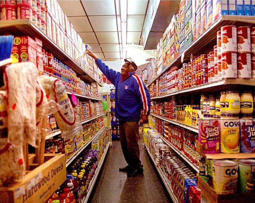 nicole gesmondi fine art photography Platts-Albany-Grocer-v2 A man in a blue jacket reaches for an item on the top shelf in a grocery store aisle lined with cans and packaged goods.