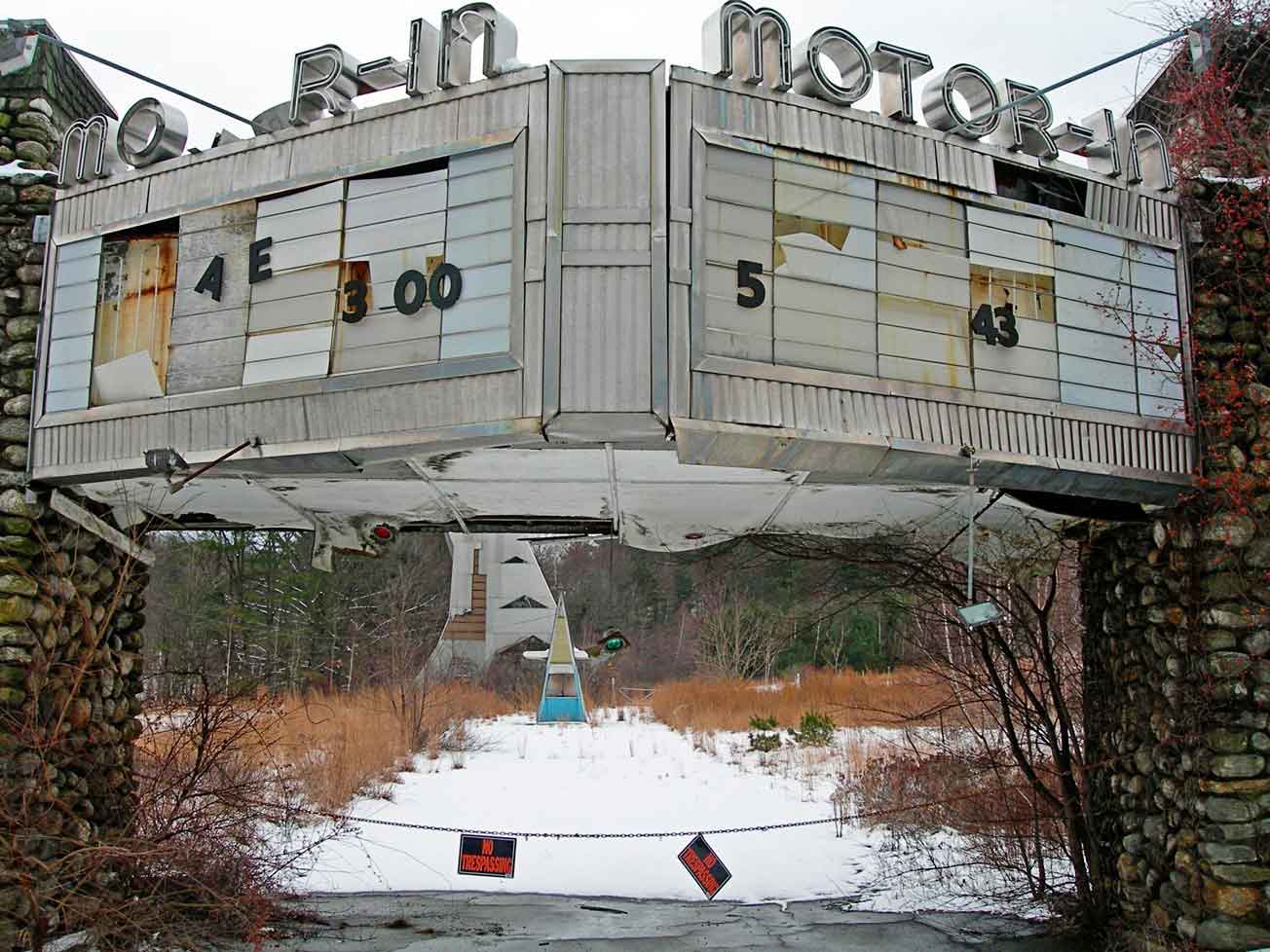 nicole gesmondi fine art photography urban x Motor-Inn An abandoned drive-in theater entrance with a weathered marquee, missing letters, overgrown weeds, and snow on the ground.