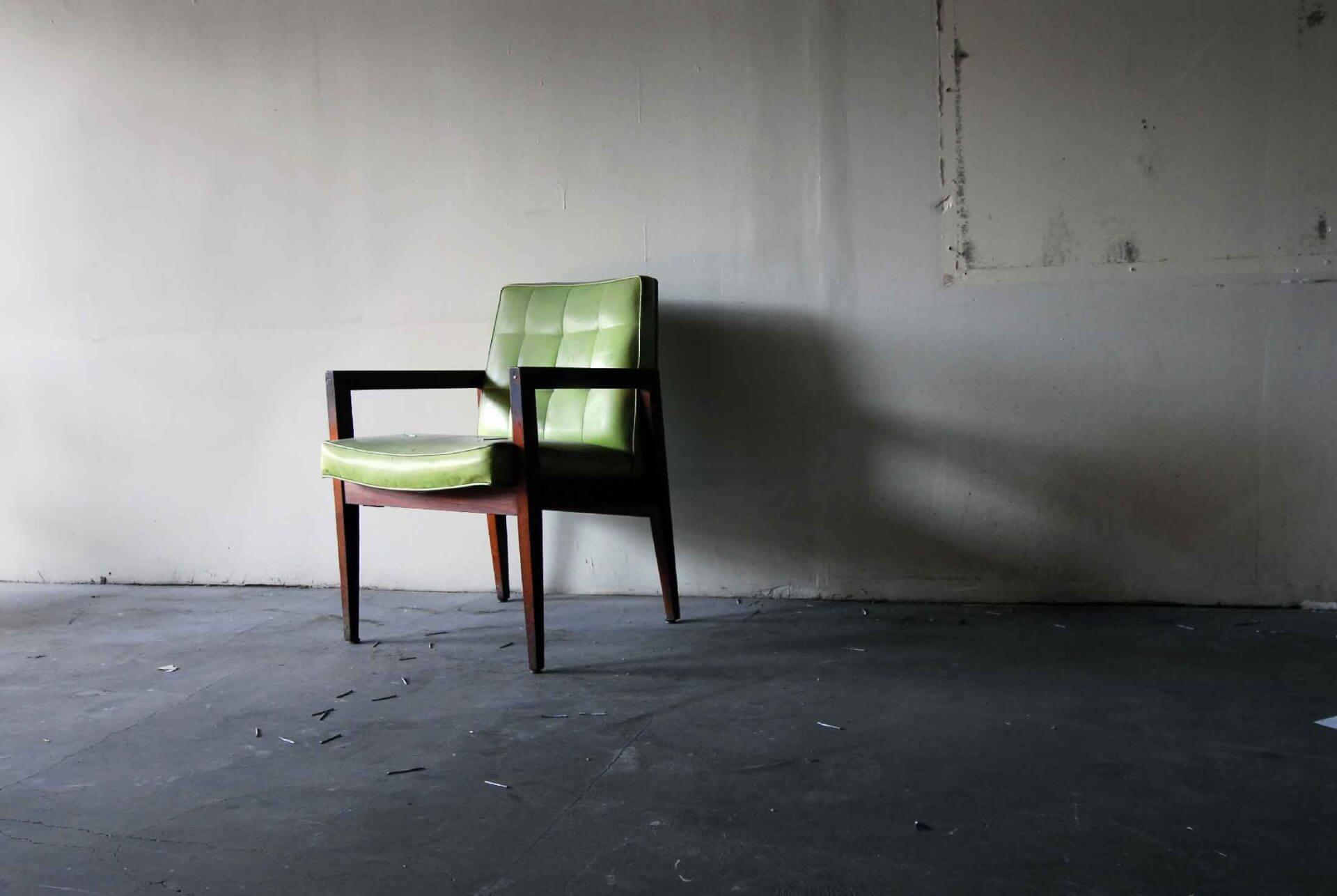nicole-gesmondi-fine-art-photography-urban-x-green-chair A single green cushioned chair with wooden arms and legs sits on a bare concrete floor against a plain, slightly marked wall.