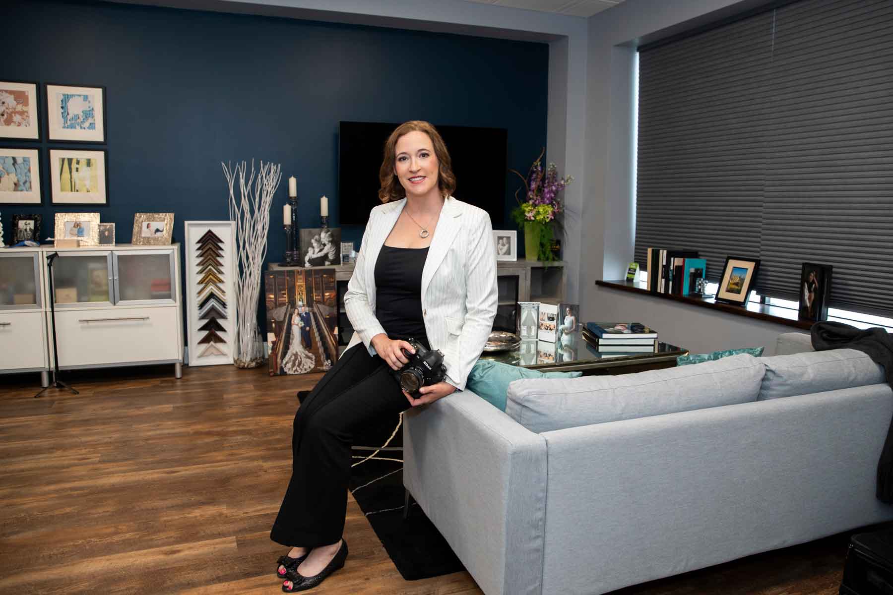 A woman in business attire sits on the armrest of a gray sofa in a modern office with blue walls, framed art, and various decor items.