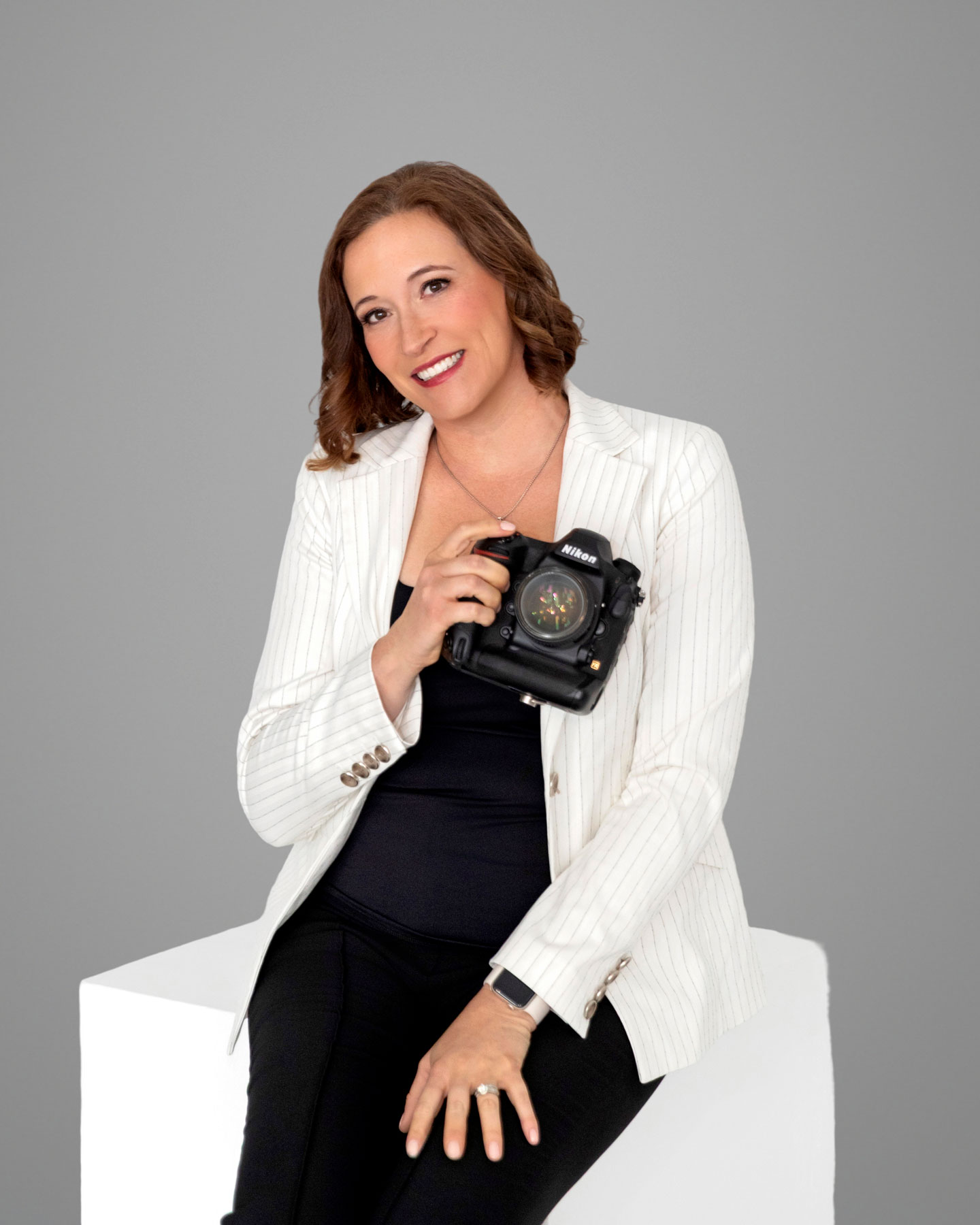 Woman in a white pinstripe blazer and black pants sits on a white block, holding a Nikon camera and smiling at the camera against a plain gray background.