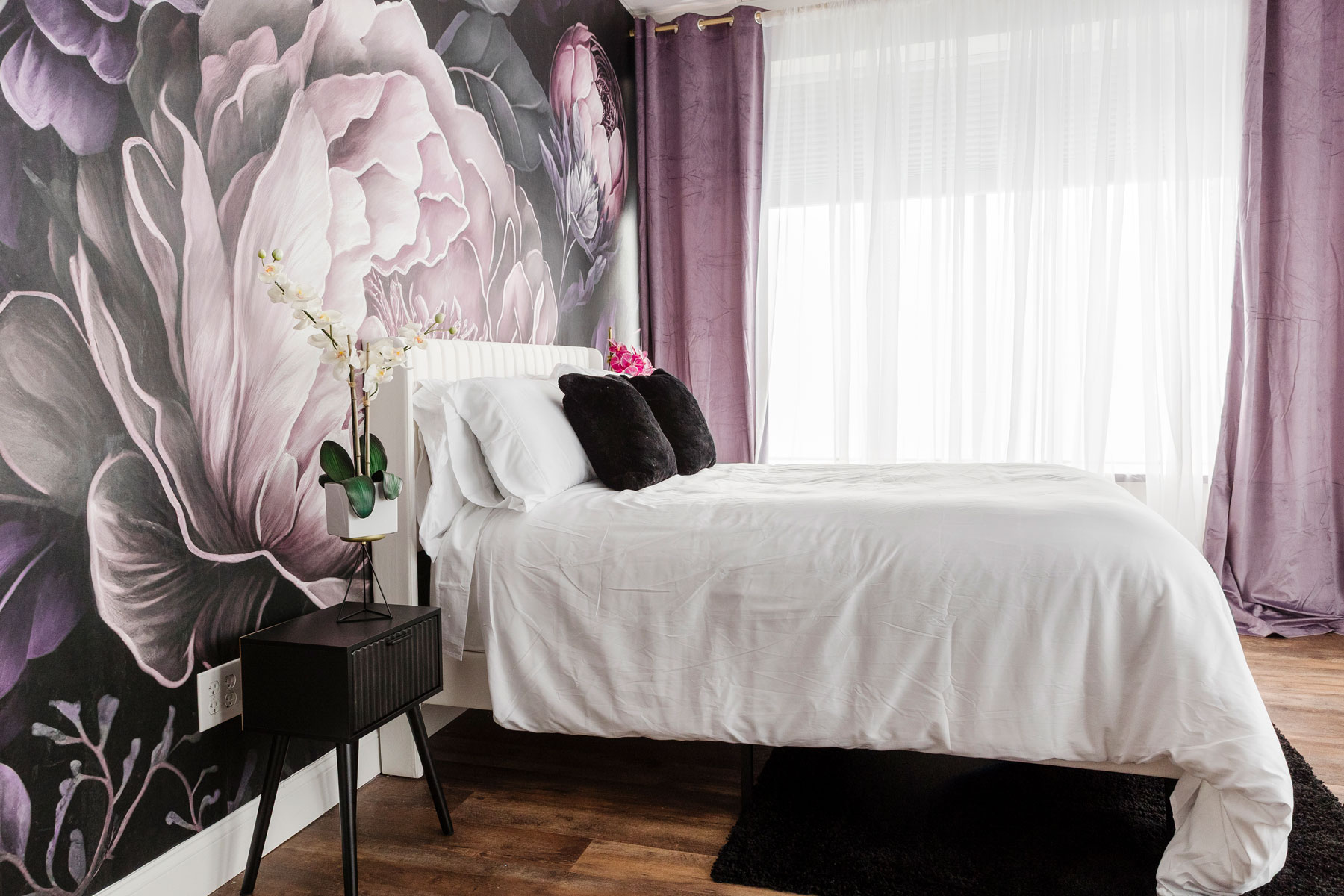 A bedroom with a white bed, black accent pillows, a floral mural on the wall, a black nightstand with a potted orchid, sheer curtains, and wood flooring.