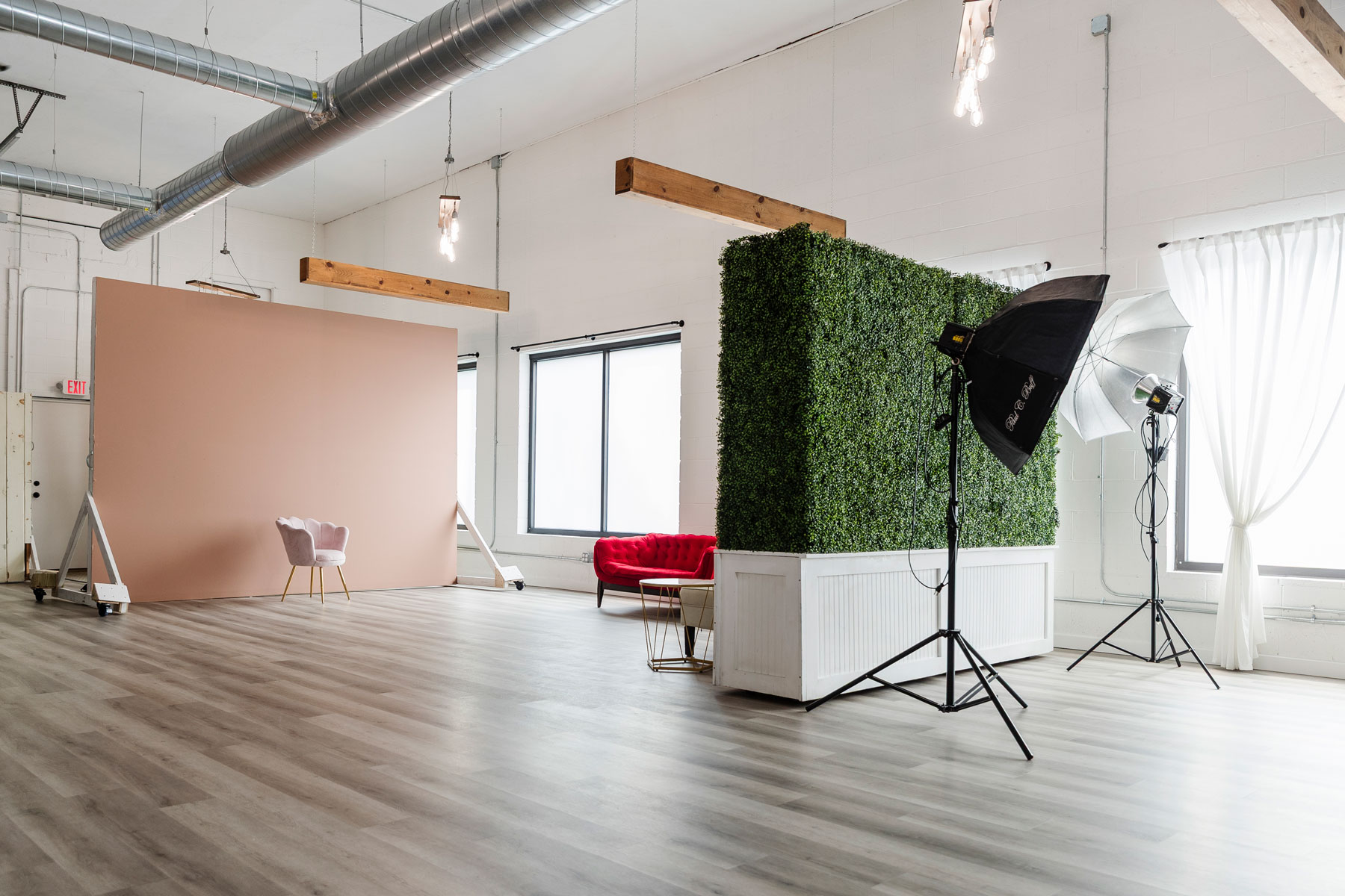 A spacious photography studio with a pink backdrop, a pink chair, a red sofa, artificial greenery, lighting equipment, large windows, and wood flooring.