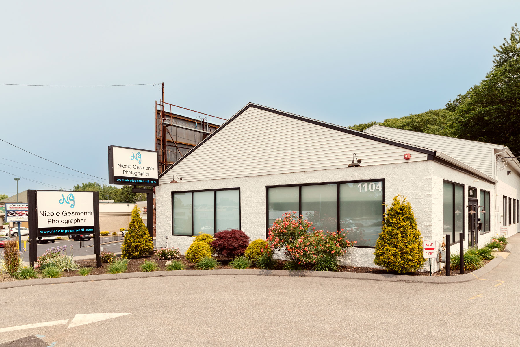 A one-story white building with large windows houses Nicole Gesmondi Photographer; signs with the business name are visible outside, and landscaped bushes line the front.