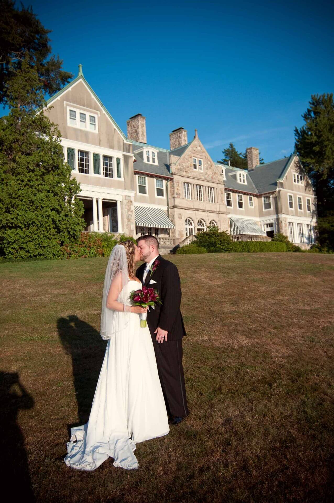 A bride and groom stand close together on a lawn in front of a large house, with the bride holding a bouquet and both facing each other under clear blue sky.