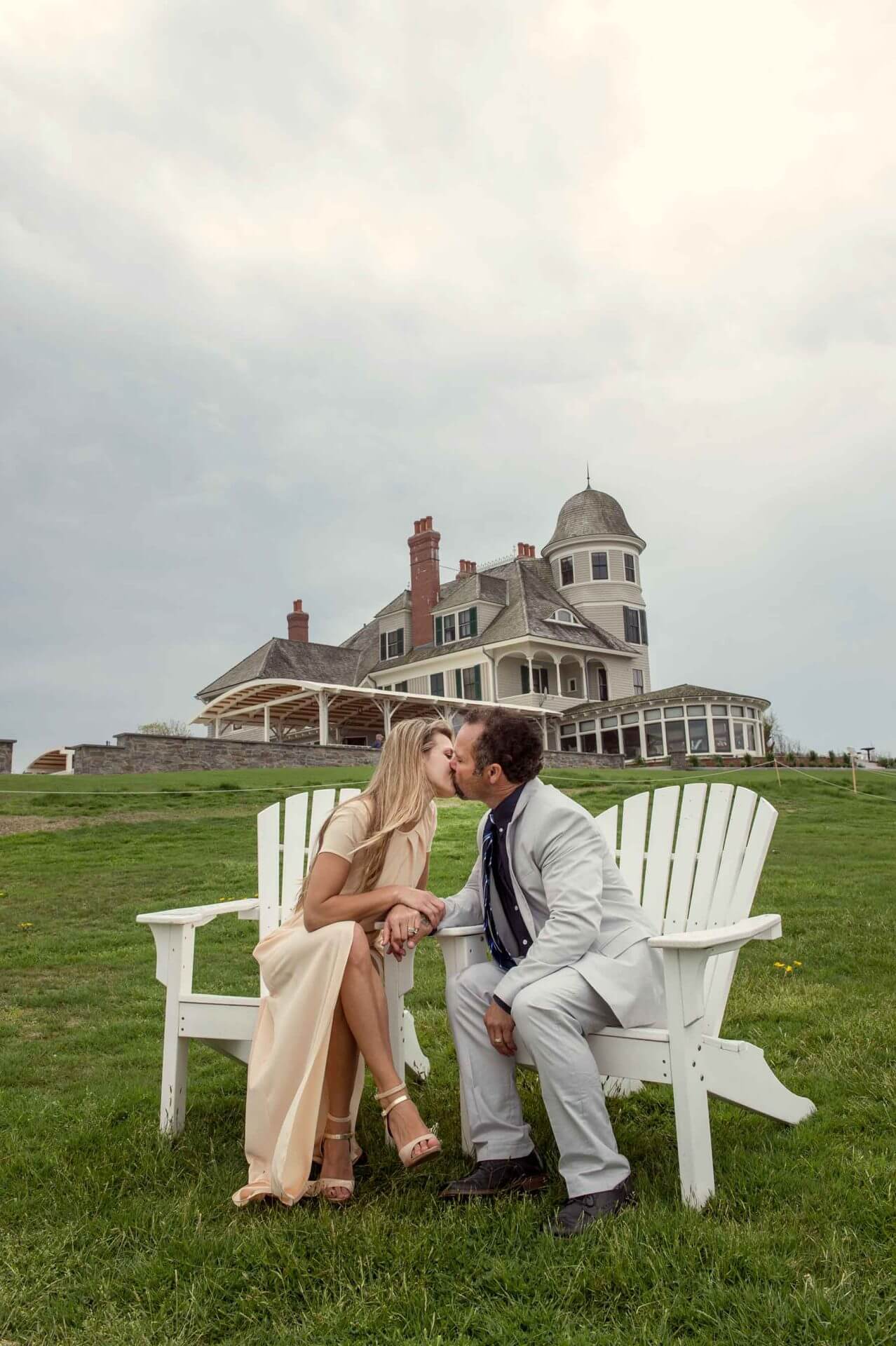 A couple dressed in formal attire sits on white chairs in front of a large historic house, sharing a kiss on a grassy lawn under a cloudy sky.