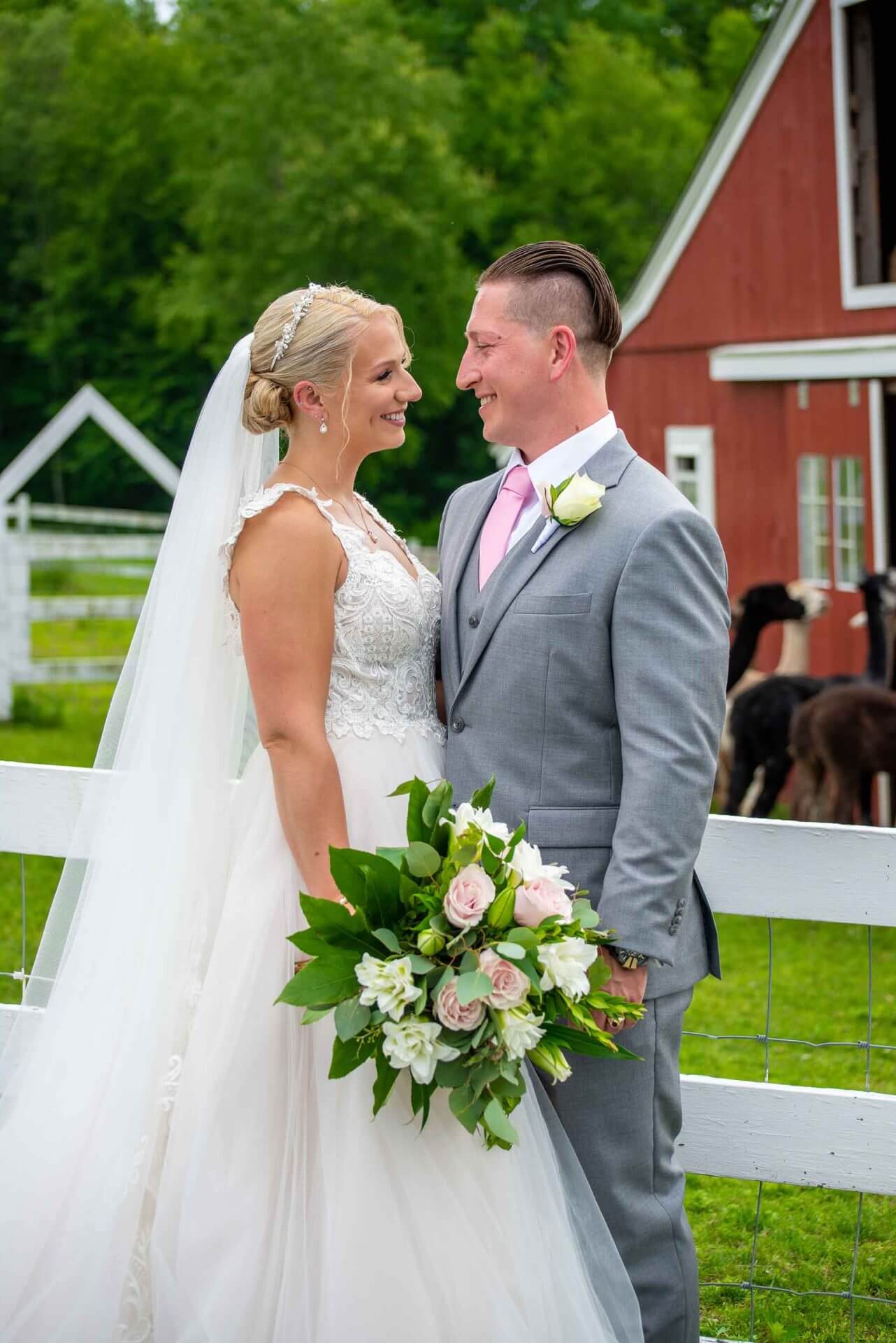 Bride and groom stand smiling at each other outdoors by a white fence, holding a bouquet, with a red barn and horses in the background.