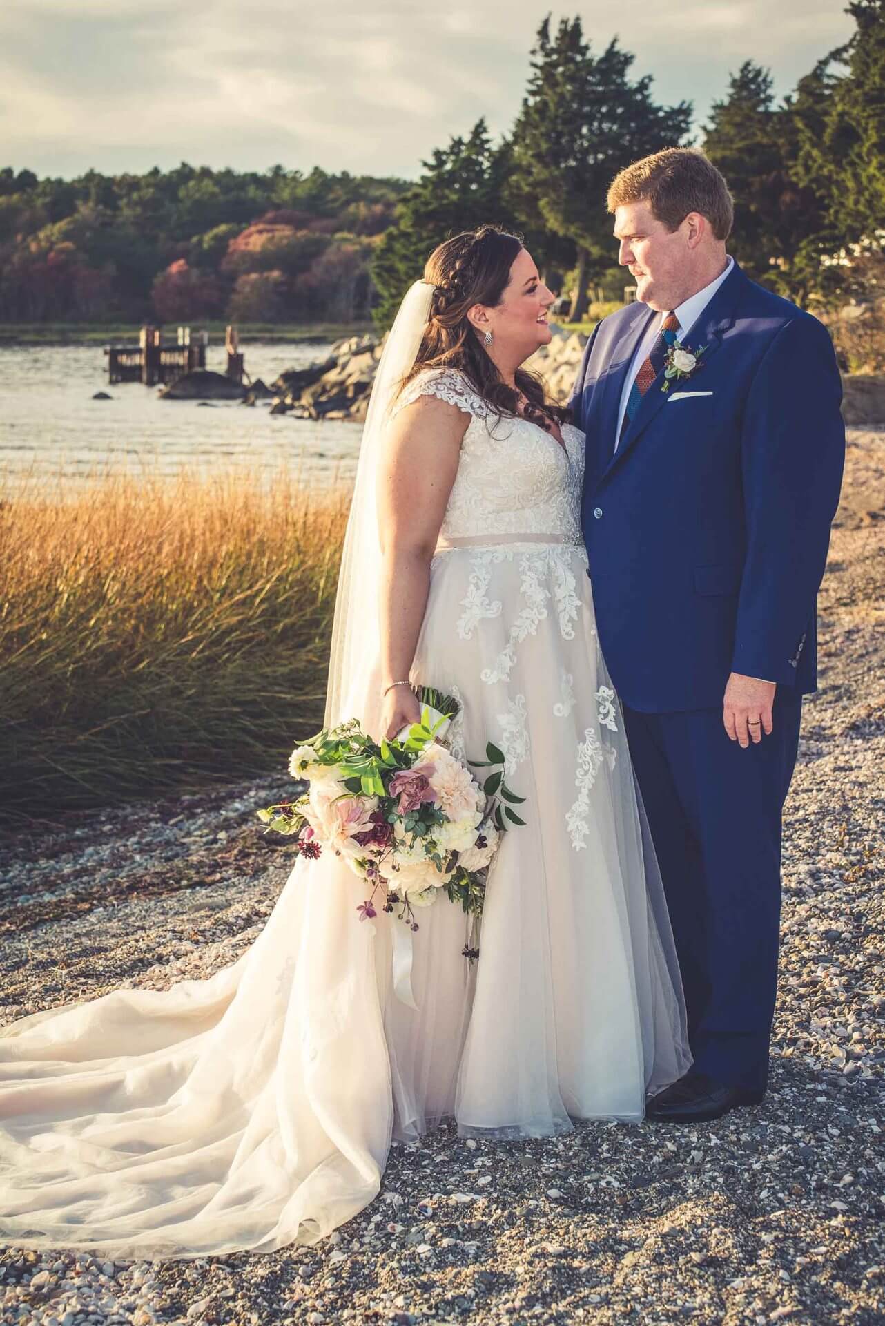 A bride and groom stand on a rocky shoreline, facing each other and smiling, with water, trees, and a dock in the background. The bride holds a bouquet of flowers.