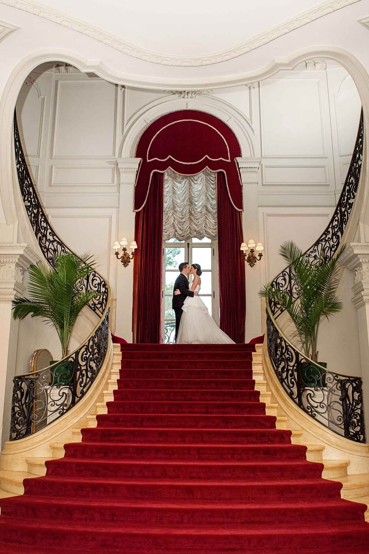 A bride and groom stand at the top of a grand staircase with red carpet, ornate railings, and elegant décor, sharing a kiss.