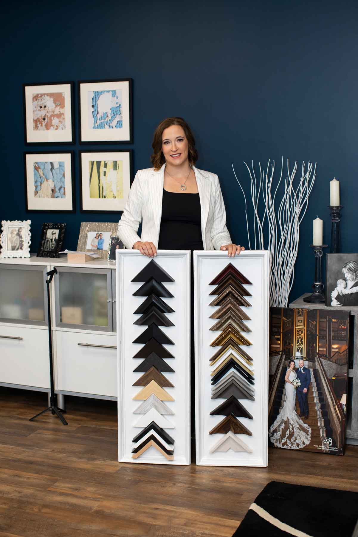 A woman stands indoors holding two display boards with various picture frame corner samples, surrounded by framed photos and decorative items.