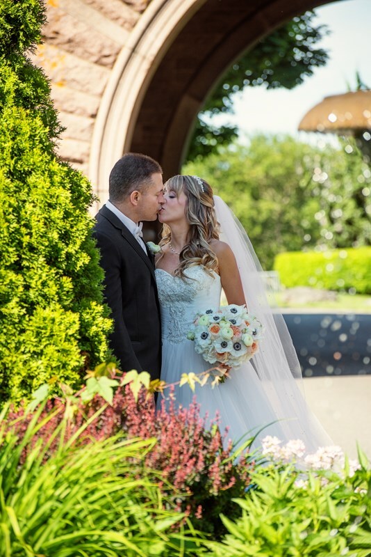 ocean-cliff-wedding-ri-028 A bride and groom in wedding attire share a kiss outdoors near greenery and flowers, with a stone arch and fountain in the background.