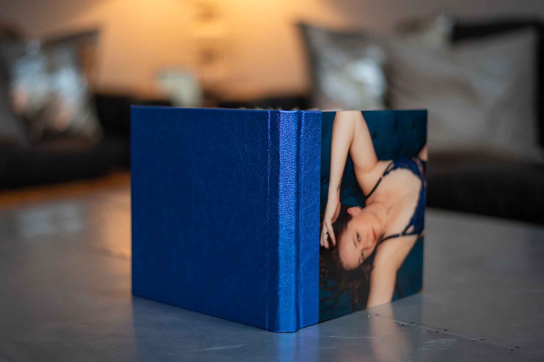 A closed photo book with a blue spine and a cover image of a woman in lingerie lying down, placed on a reflective surface with a blurred background.