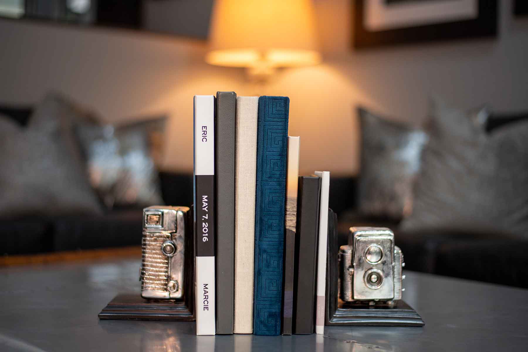 Books are positioned upright between two vintage camera bookends on a coffee table, with a lit lamp and sofa in the blurred background.