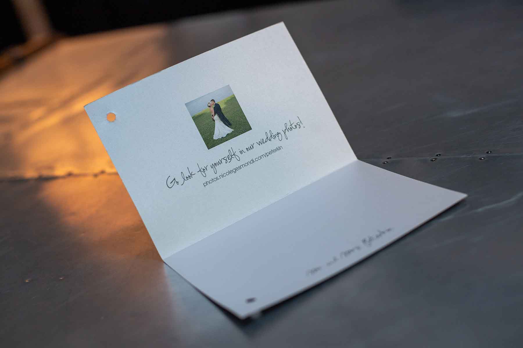 A wedding invitation card with a small photo of a couple and text inviting guests to view wedding photos, placed on a metallic surface.
