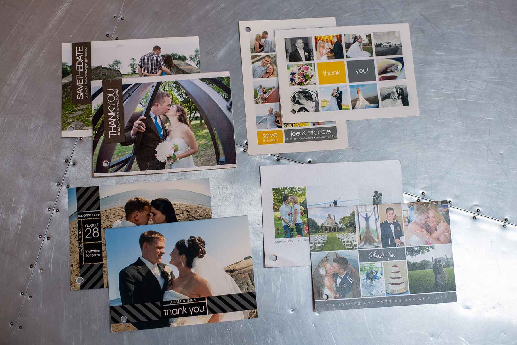 A variety of wedding invitation and thank you cards featuring photos of couples and decorative text are arranged on a metallic surface.