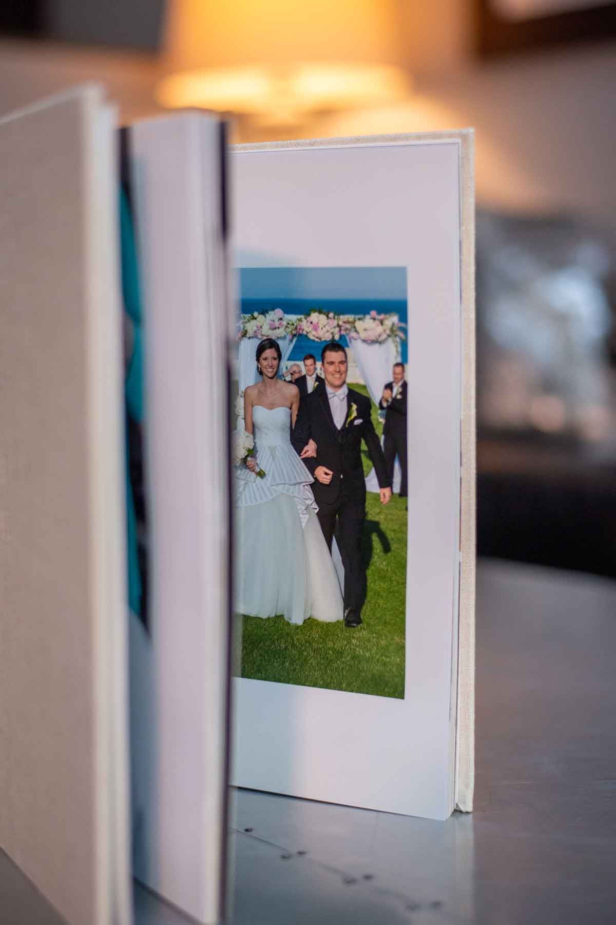 print-products-wedding-album-interior-image An open photo album displays a wedding photo of a bride and groom walking outdoors under a decorated arch.