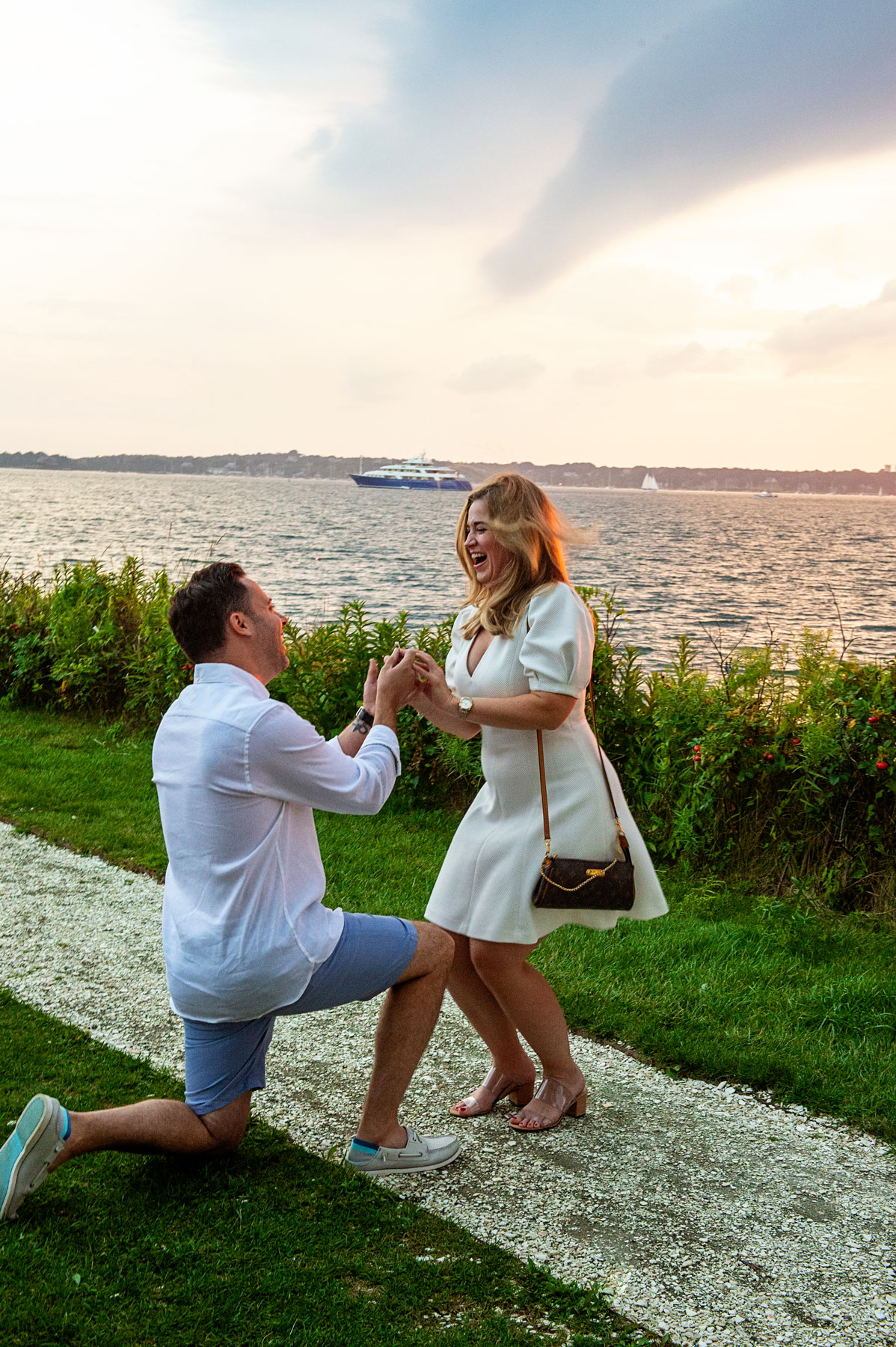 A man kneels and proposes to a woman by the water at sunset. She looks surprised and happy, holding his hands. A boat is visible in the background.