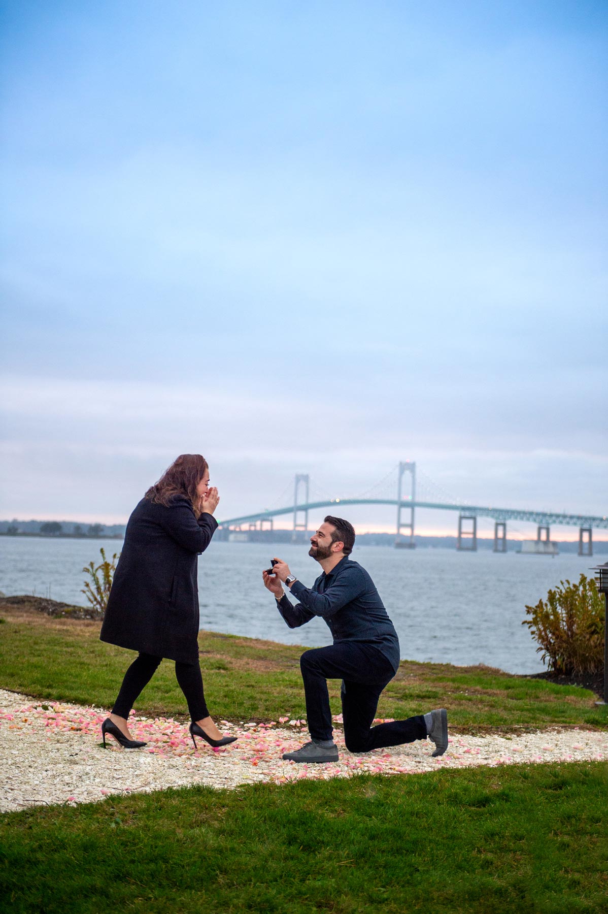A man kneels and holds a ring box, proposing to a woman outdoors near water with a bridge in the background. The woman appears surprised and joyful.