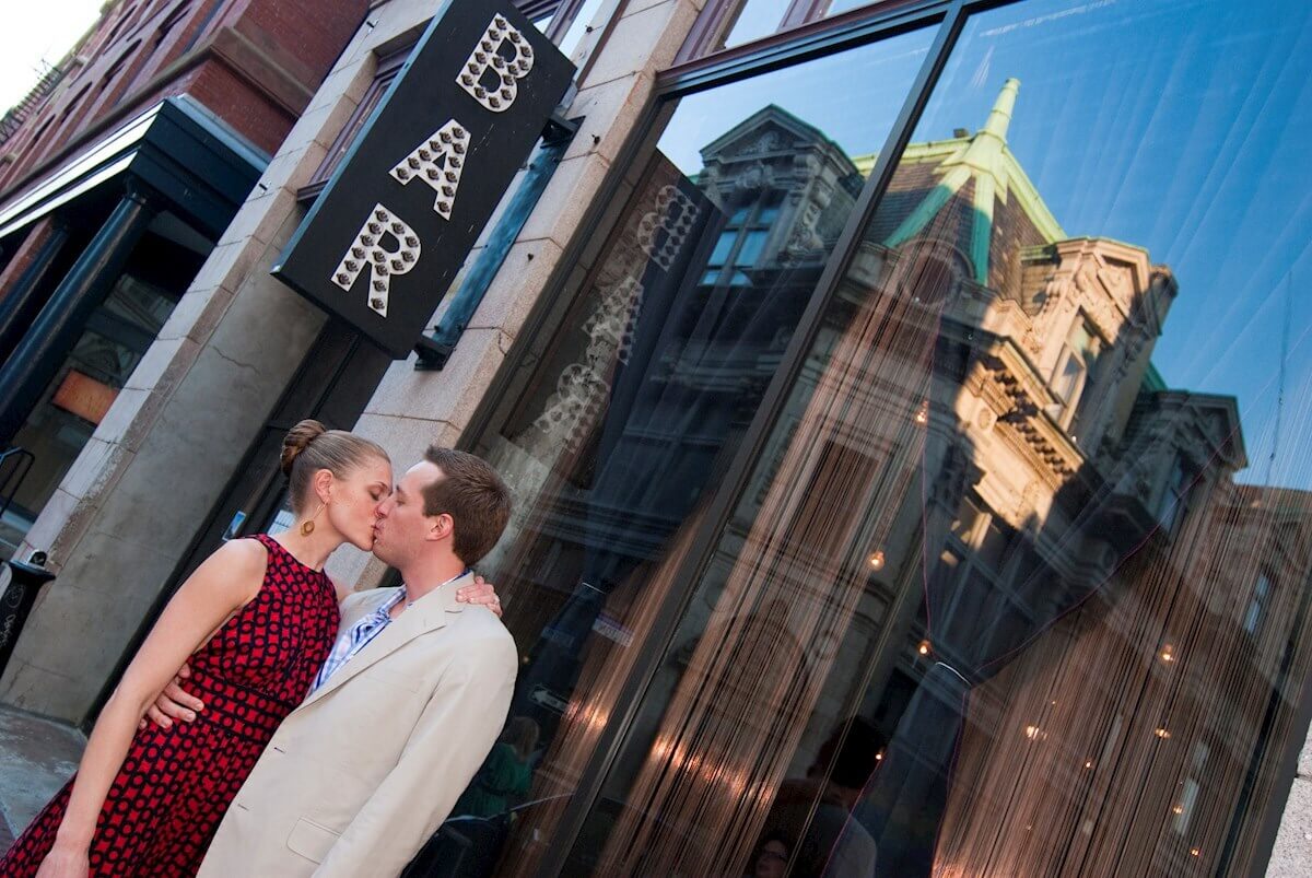 ri-rehearsal-dinner-photographer-001 A couple kisses outside a bar with a large sign, with city buildings reflected in the glass windows.