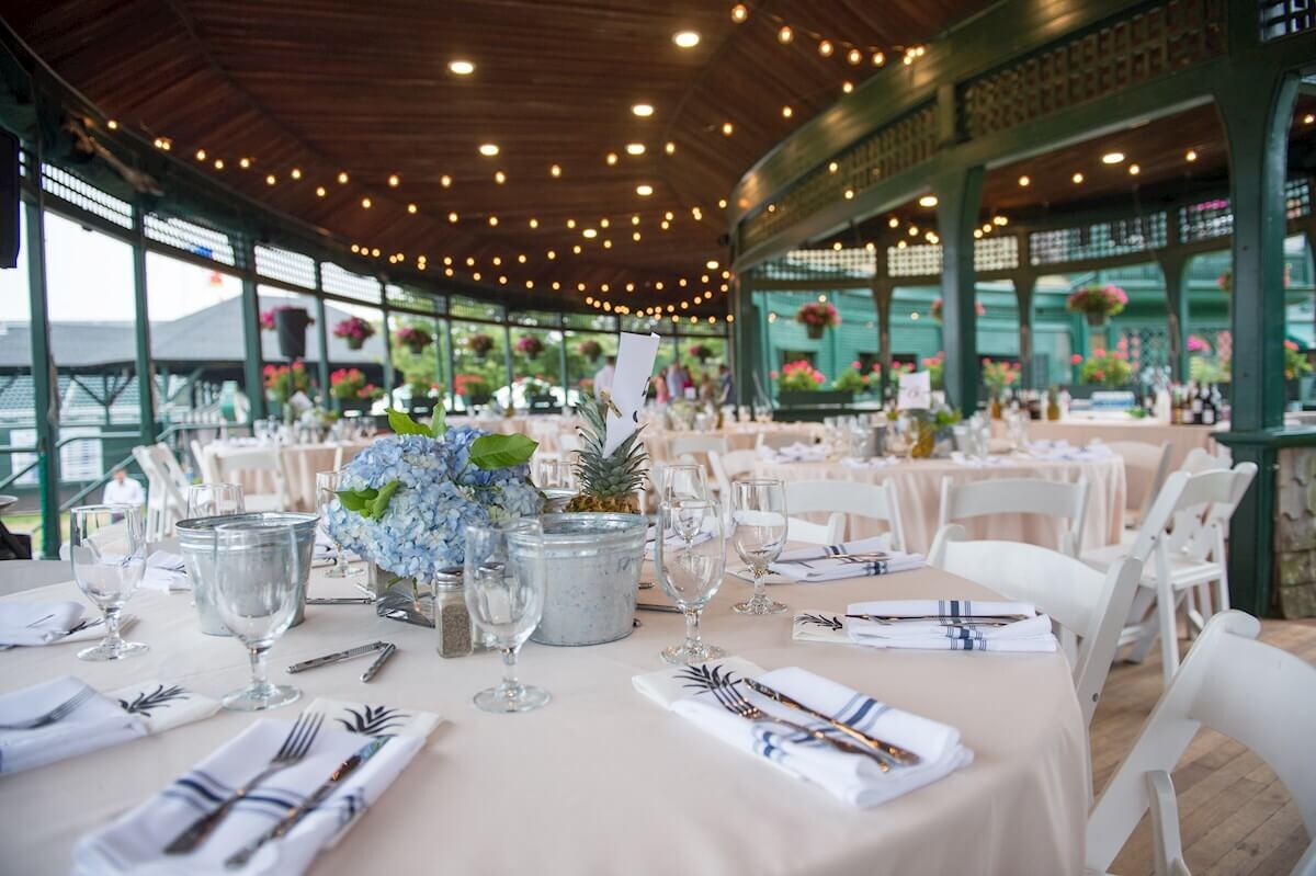 ri-rehearsal-dinner-photographer-002 Round tables with white tablecloths are set for an event under a wooden pavilion with string lights, floral centerpieces, and neatly arranged place settings.