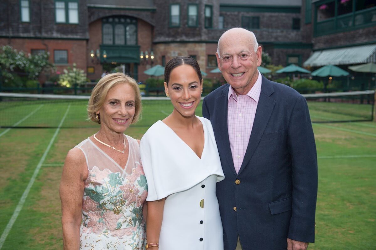 ri-rehearsal-dinner-photographer-003 Three people stand and smile on a grass tennis court with a building and umbrellas in the background.
