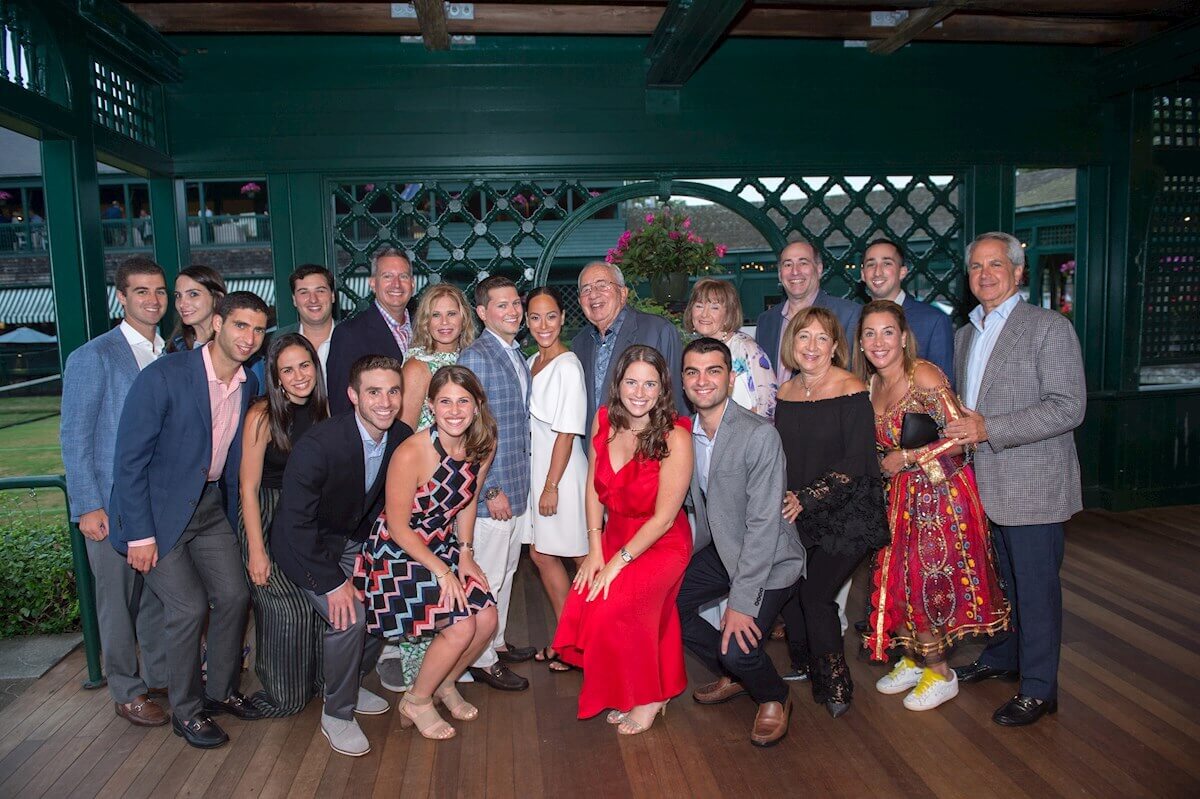 ri-rehearsal-dinner-photographer-004 A group of about twenty people, dressed in semi-formal and formal attire, pose together and smile for a photo on a covered outdoor wooden deck.