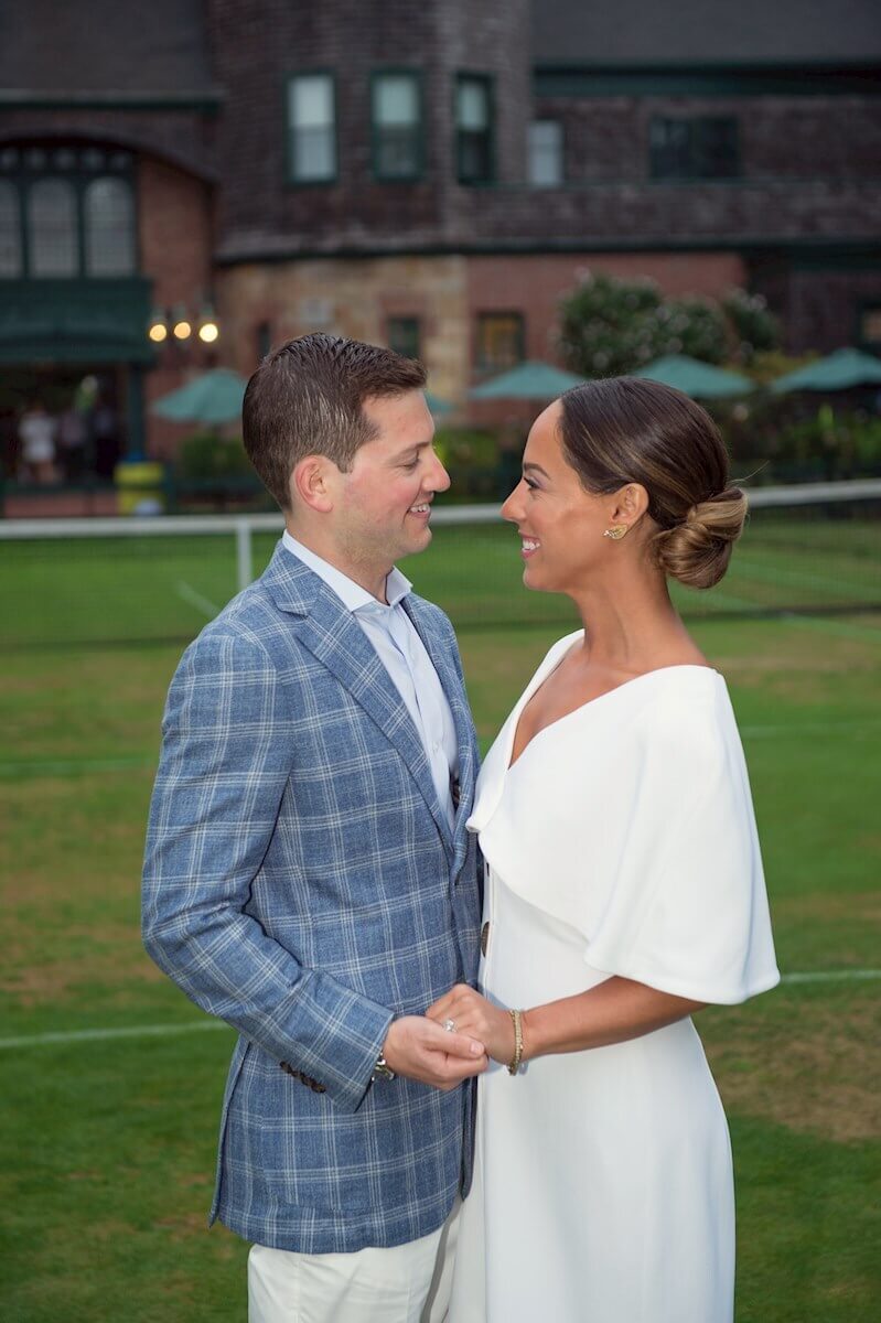 ri-rehearsal-dinner-photographer-005 A man and woman dressed formally stand outdoors on grass, holding hands and smiling at each other with a large building in the background.