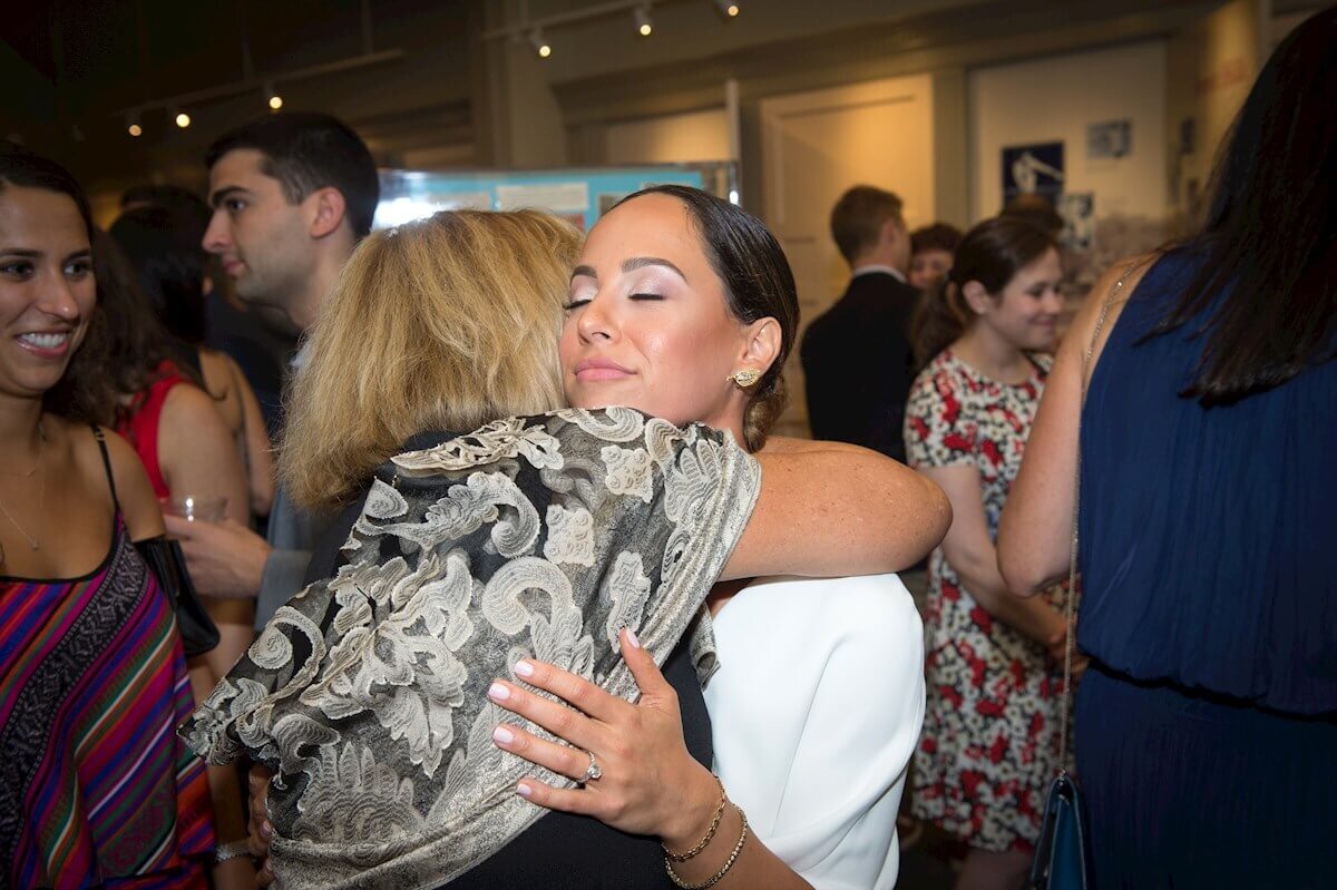 ri-rehearsal-dinner-photographer-009 Two women embrace in a hug at an indoor social gathering, surrounded by other people who are talking and mingling.