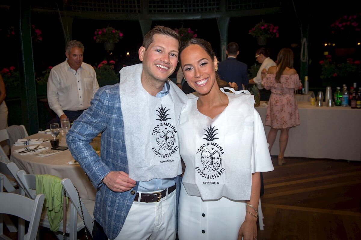 ri-rehearsal-dinner-photographer-010 A smiling man and woman wearing custom bibs pose together at an indoor event with other guests and a buffet table in the background.