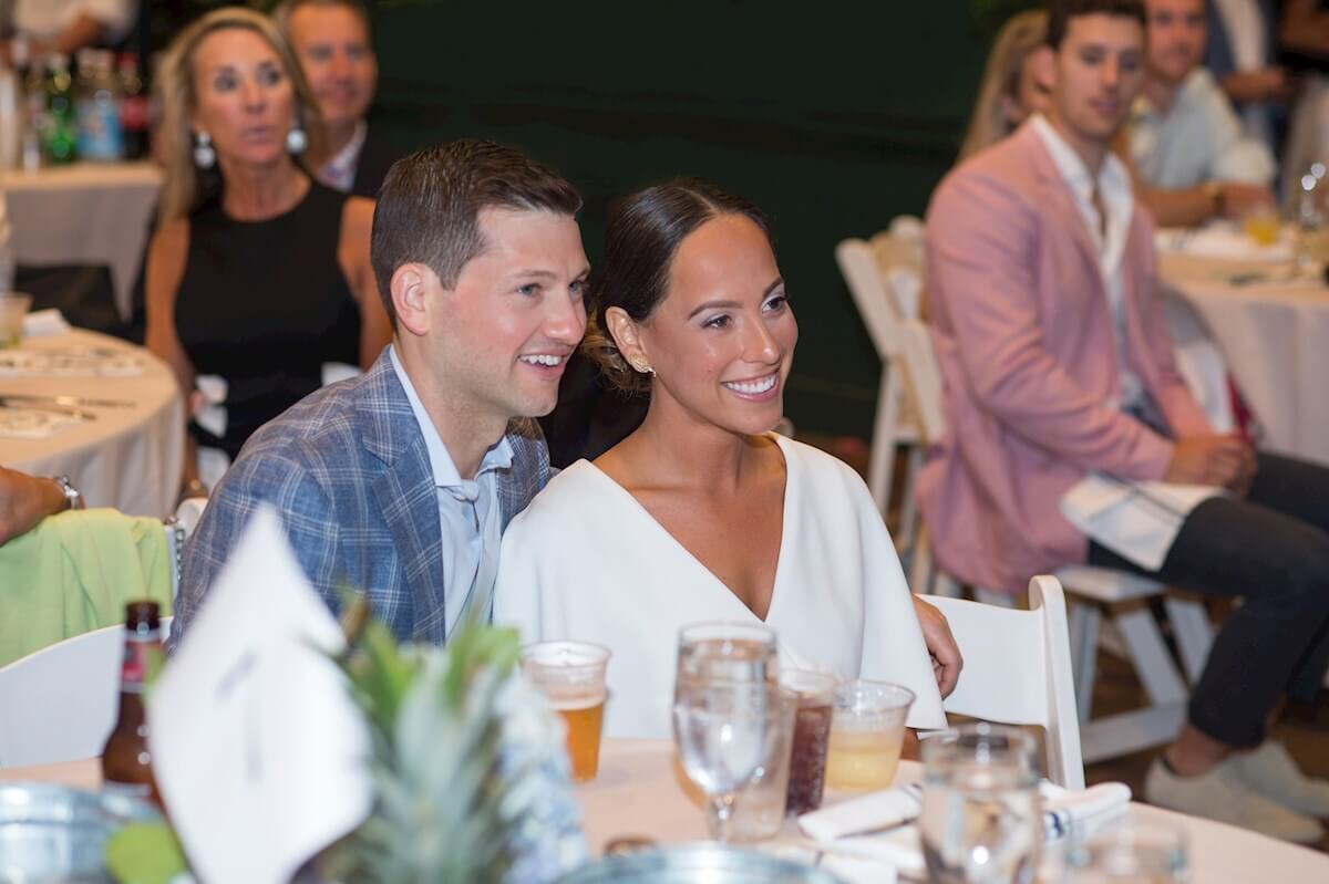 ri-rehearsal-dinner-photographer-011 A man and woman sit closely together, smiling at an event with tables, drinks, and other attendees visible in the background.