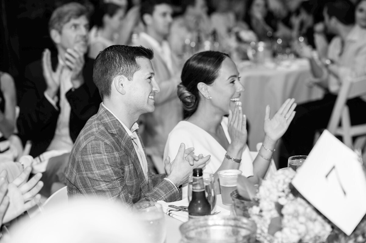 ri-rehearsal-dinner-photographer-012 A man and woman sit at a table, smiling and applauding, surrounded by other people doing the same at a formal event.