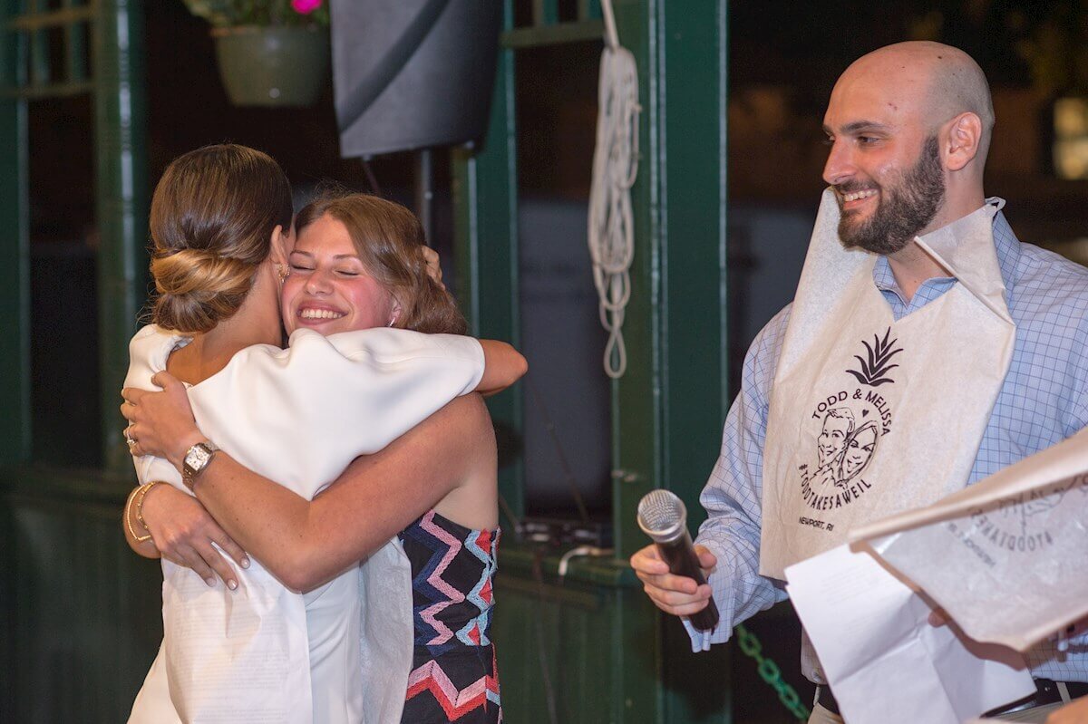 ri-rehearsal-dinner-photographer-013 Two women embrace and smile while a man holding a microphone and papers stands nearby, wearing a napkin bib at an indoor event.