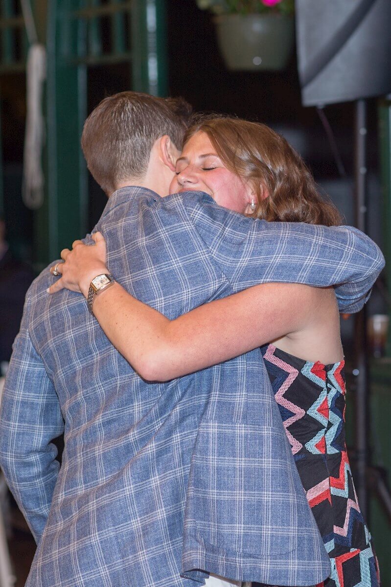 ri-rehearsal-dinner-photographer-014 Two people embrace in a hug; one wears a plaid jacket, the other a geometric-patterned dress. Both appear to be smiling. Indoor setting with blurred background elements.