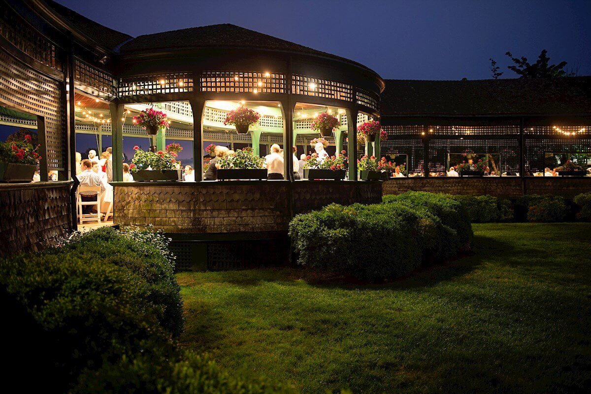 ri-rehearsal-dinner-photographer-016 An outdoor pavilion with people dining under warm lights at dusk, surrounded by green shrubs and hanging flower baskets.