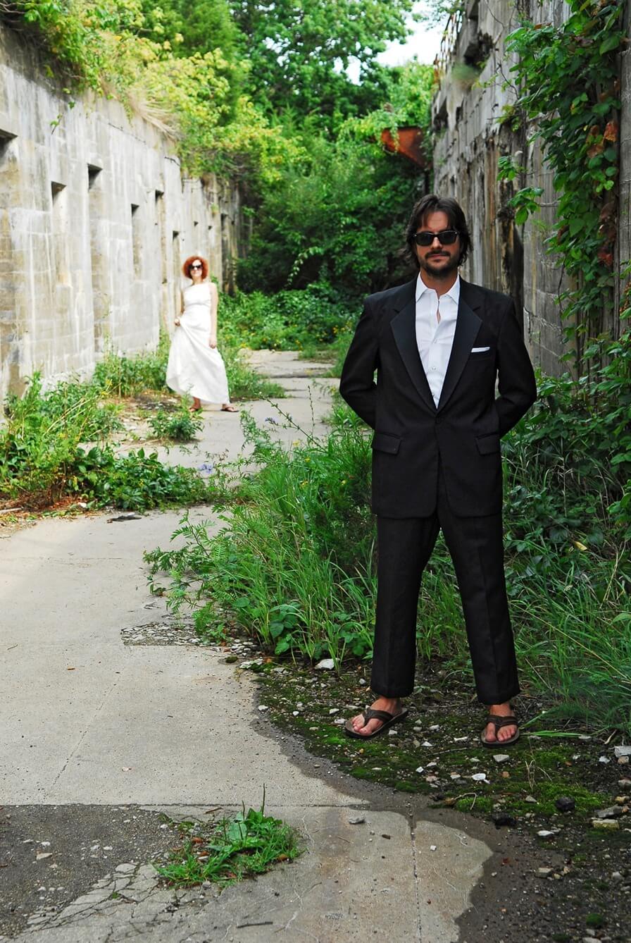 ri-trash-the-dress-photographer-001 A man in a black suit and sunglasses stands on a path with overgrown vegetation; a woman in a white dress stands further back.