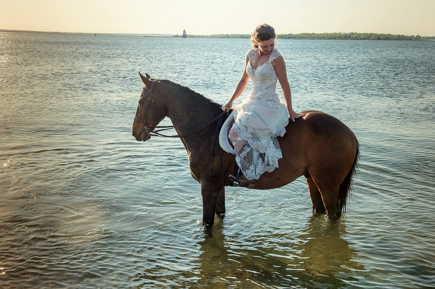 ri-trash-the-dress-photographer-002 A woman in a white dress sits on a brown horse standing in shallow water near a shoreline on a sunny day.