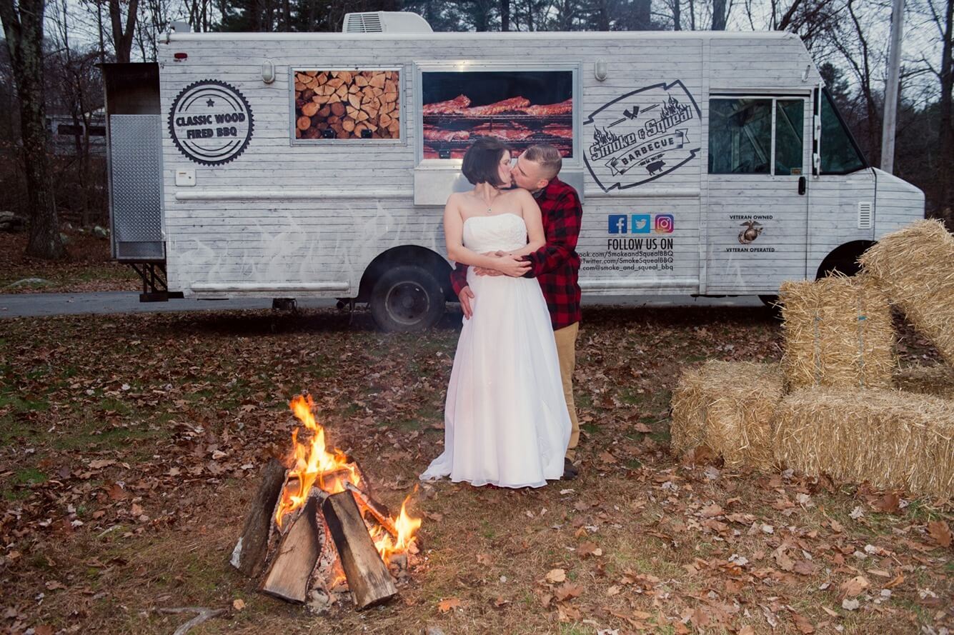 ri-trash-the-dress-photographer-004 A couple in wedding attire kisses in front of a lit campfire, with a food truck and hay bales in the background, set outdoors on fallen leaves.